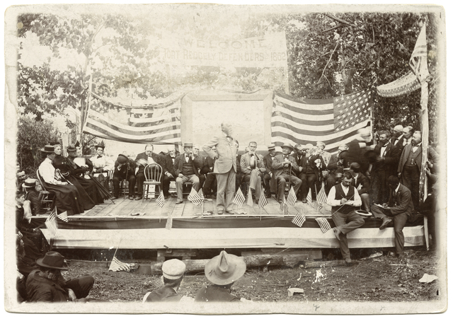 Stage platform with observers in front and people sitting in chairs in front of them, with a speaker standing front and center.